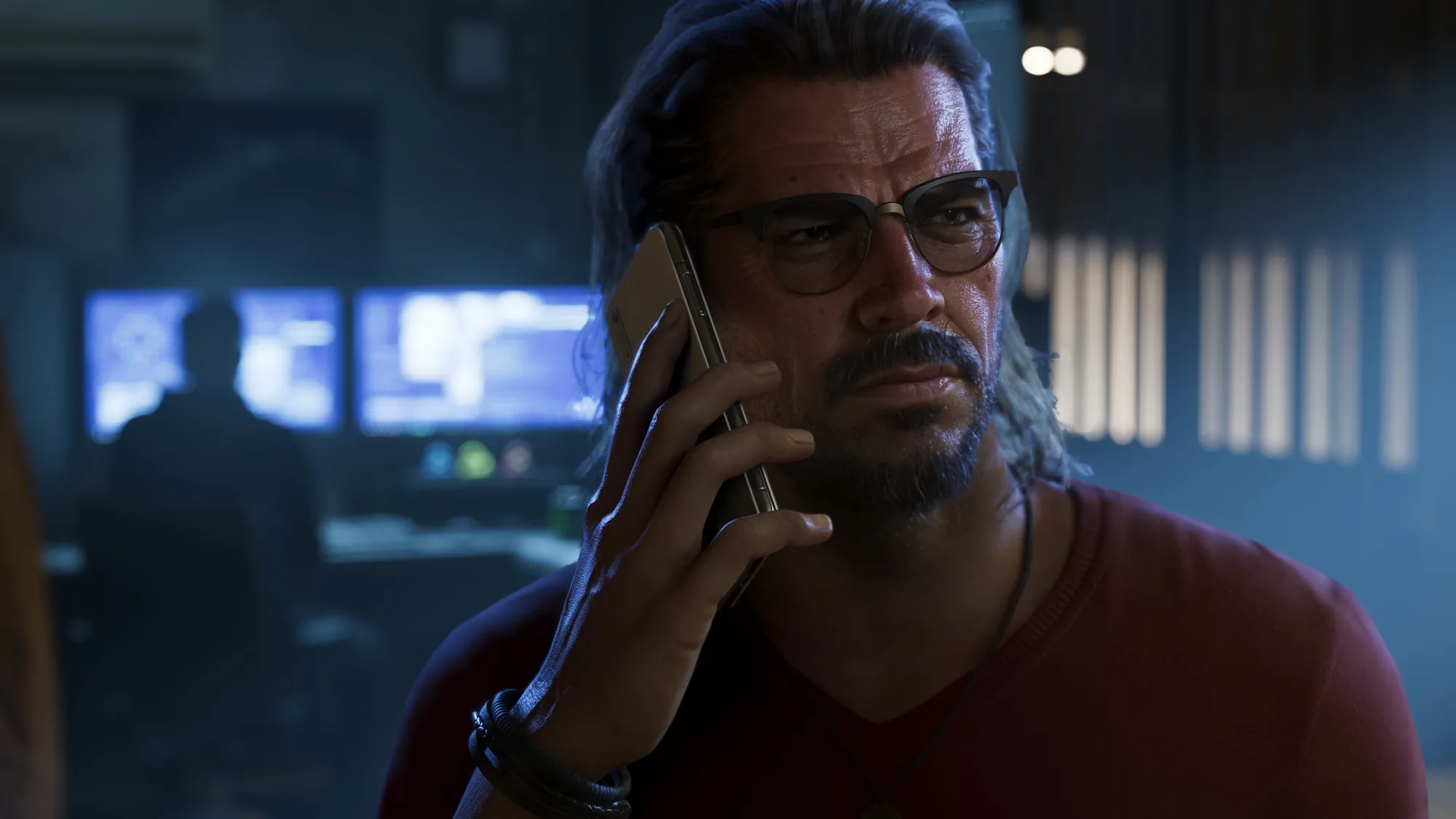 A bearded man with shoulder-length grey hair and glasses talks on a phone in a neon-lit interior at night, wearing a red shirt.